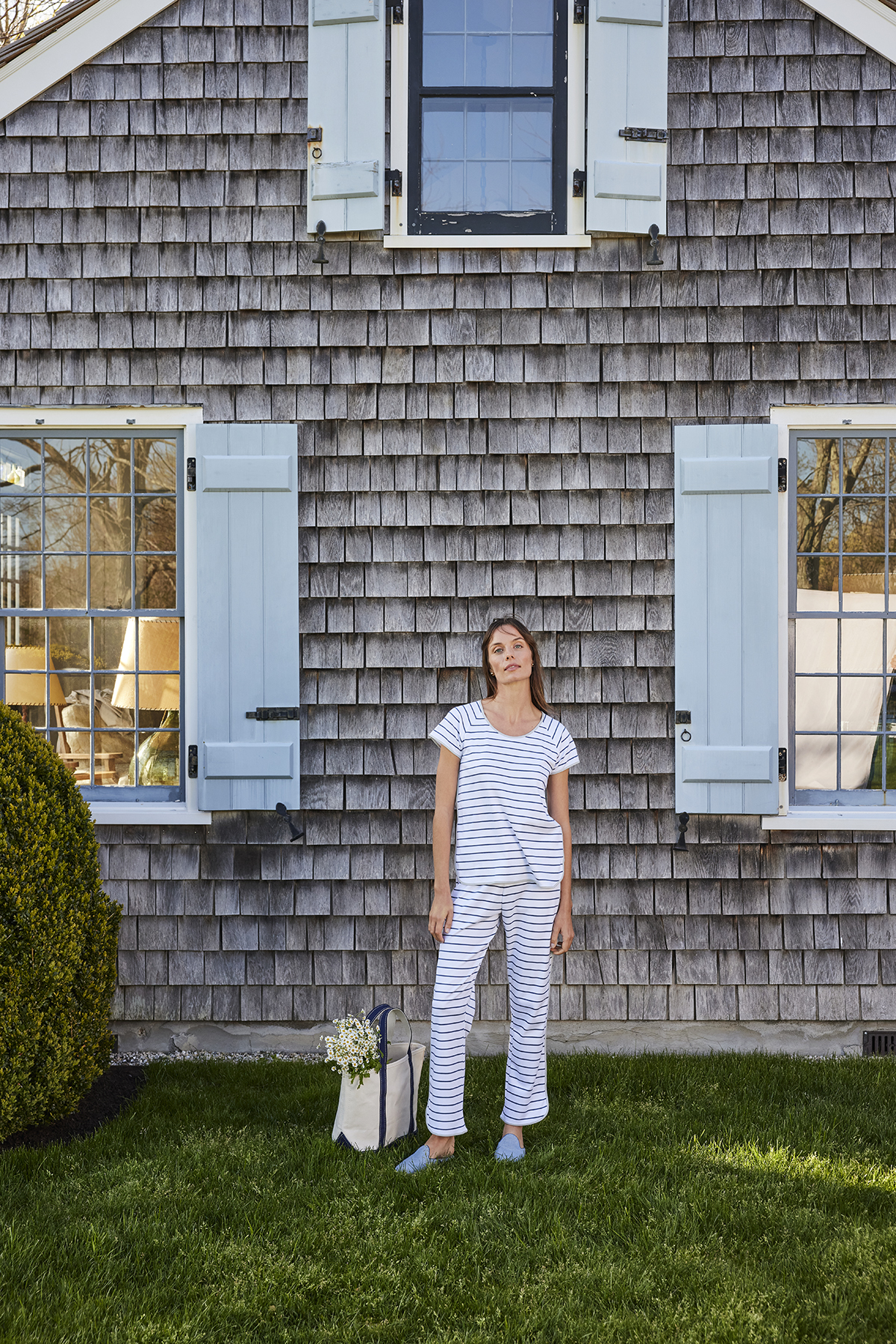 Woman in striped pajamas stands in front of wood-shingled house with blue shutters on green lawn holding flowers and tote bag.