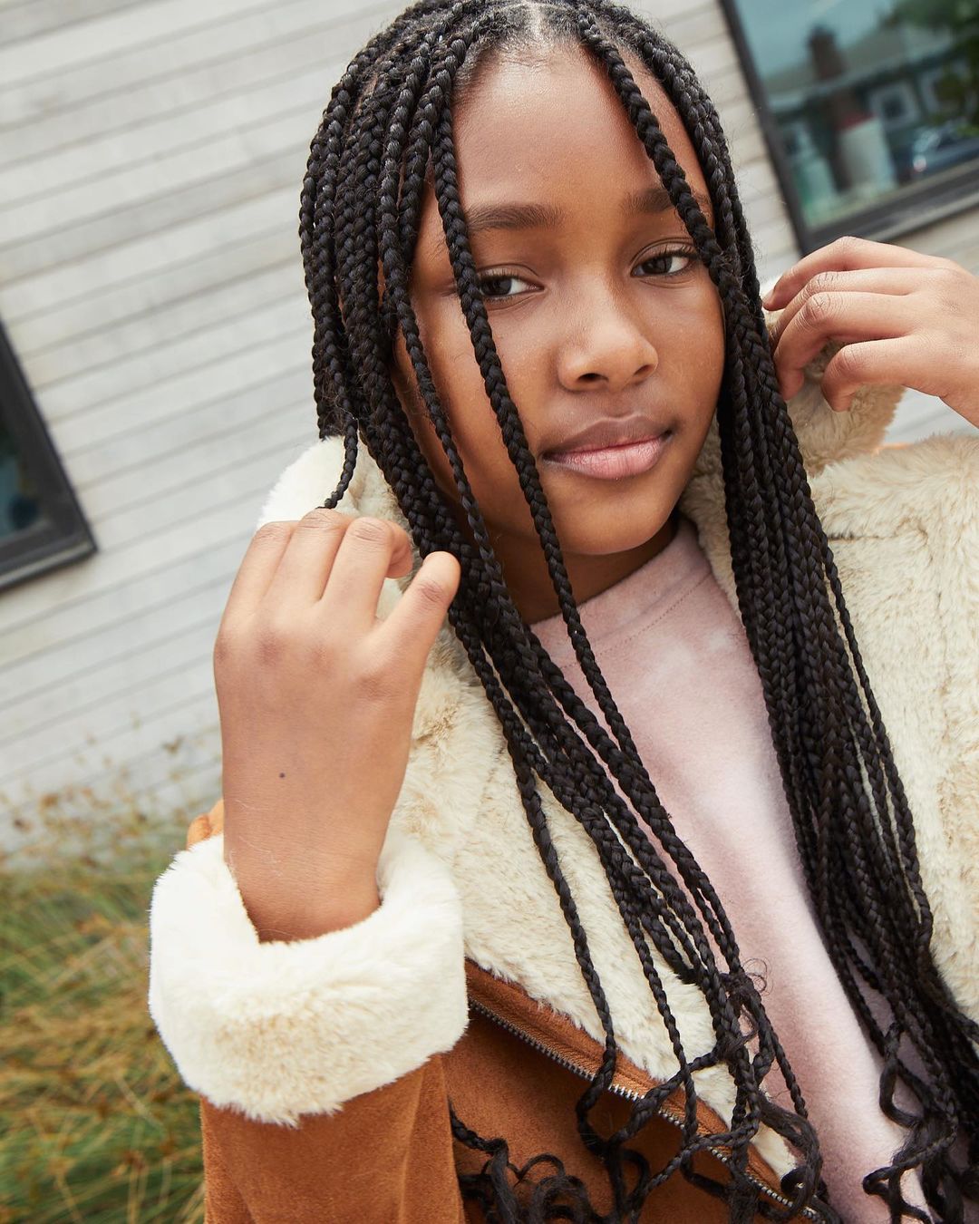 Portrait of a young Black female with braids wearing a shearling-lined brown jacket. Focus on her face and upper body in an outdoor setting.