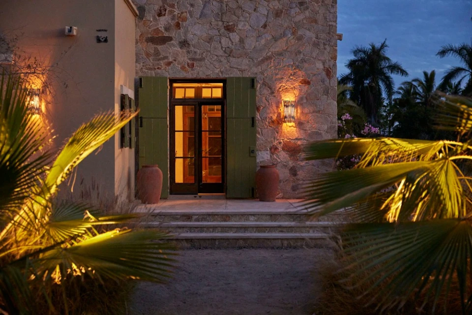Exterior view of a stone building with warm lighting, showcasing a dark wood door, green shutters, and clay pots in a tropical setting.