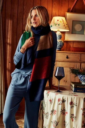 Woman wearing blue loungewear and a multi-colored striped scarf standing indoors by a table with a wine glass.