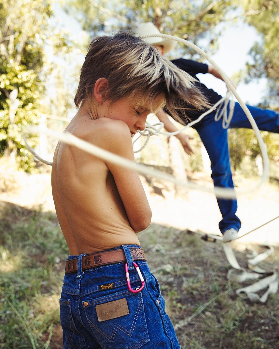 A shirtless young boy stands outdoors wearing jeans with a rope around him, while another child plays with a rope in the background.