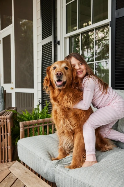 A young girl hugs a golden retriever dog on a cushioned outdoor sofa, with a porch and house in the background, creating a heartwarming scene.