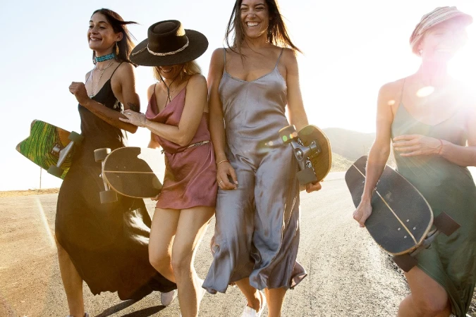 Four smiling women walking along an outdoor paved road while holding skateboards under golden hour sunlight.