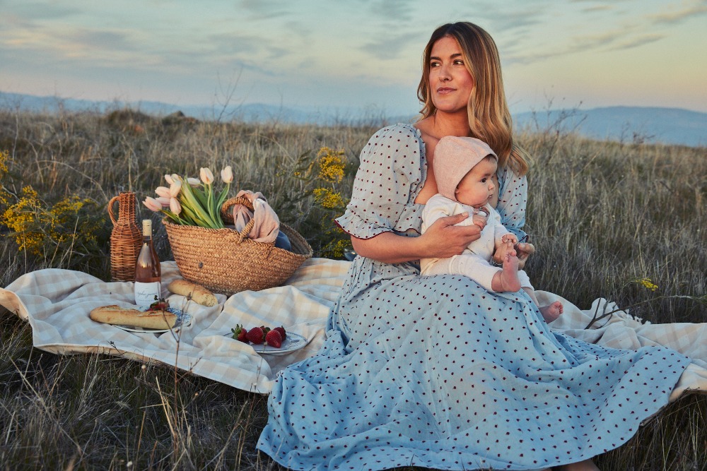 Woman holds baby on blanket in field at picnic with wine, strawberries, flowers, bread. Mountains visible in the soft sunset light.