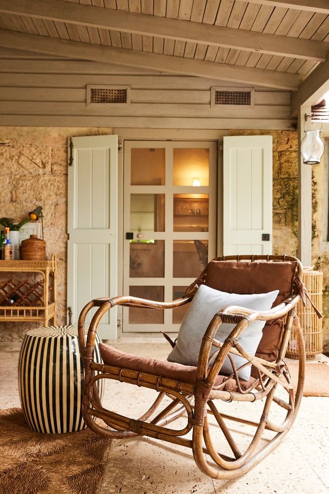 Wicker rocking chair with a pillow on a stone patio with a garden stool and a bar cart in the background.