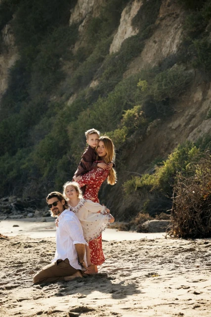 Family portrait on a beach with parents and two children, featuring a father kneeling, daughter standing on his back, mother, and son.