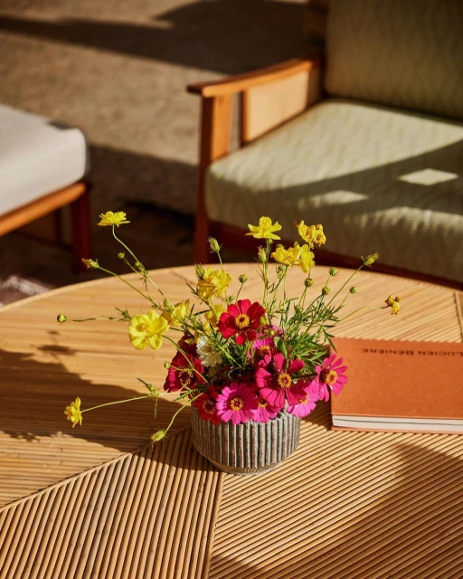 Floral arrangement of yellow and pink cosmos flowers in a textured vase on a round table next to a book and armchairs.