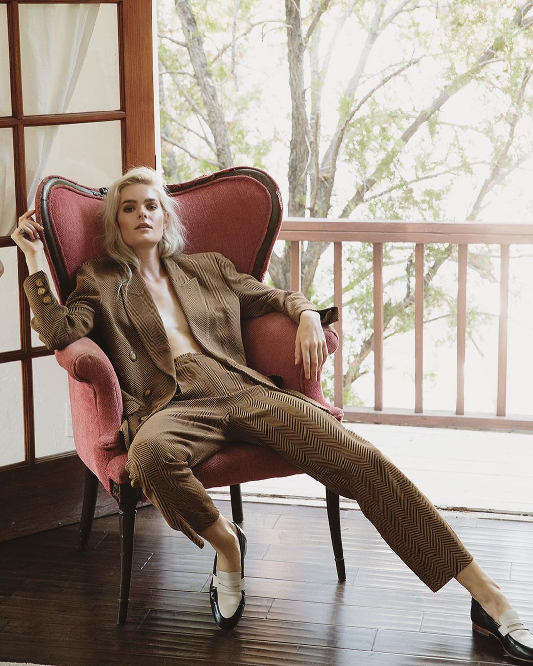 Woman in a brown pantsuit poses in a pink chair with a window background and a view of trees on a bright day.