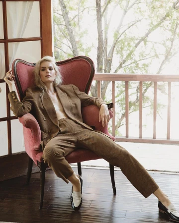Woman in a brown pantsuit poses in a pink chair with a window background and a view of trees on a bright day.