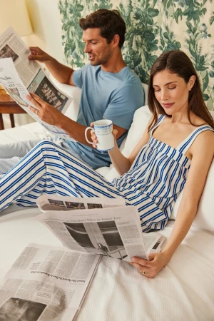 Adult couple reads newspapers in bed with floral wallpaper and white sheets. Morning sunlight illuminates relaxed home lifestyle scene.