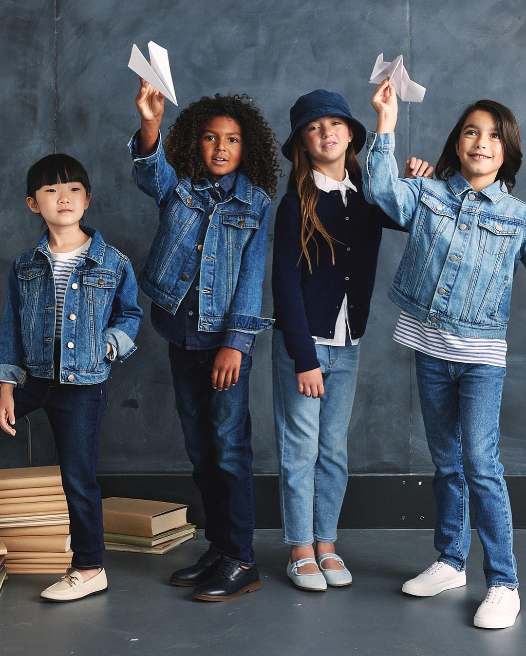 Four children stand indoors against a gray backdrop, wearing denim jackets, and holding paper airplanes, in a group studio shot.