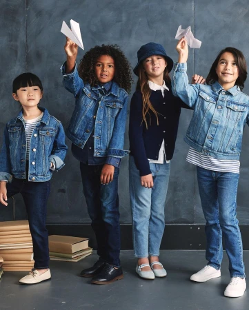 Four children stand indoors against a gray backdrop, wearing denim jackets, and holding paper airplanes, in a group studio shot.