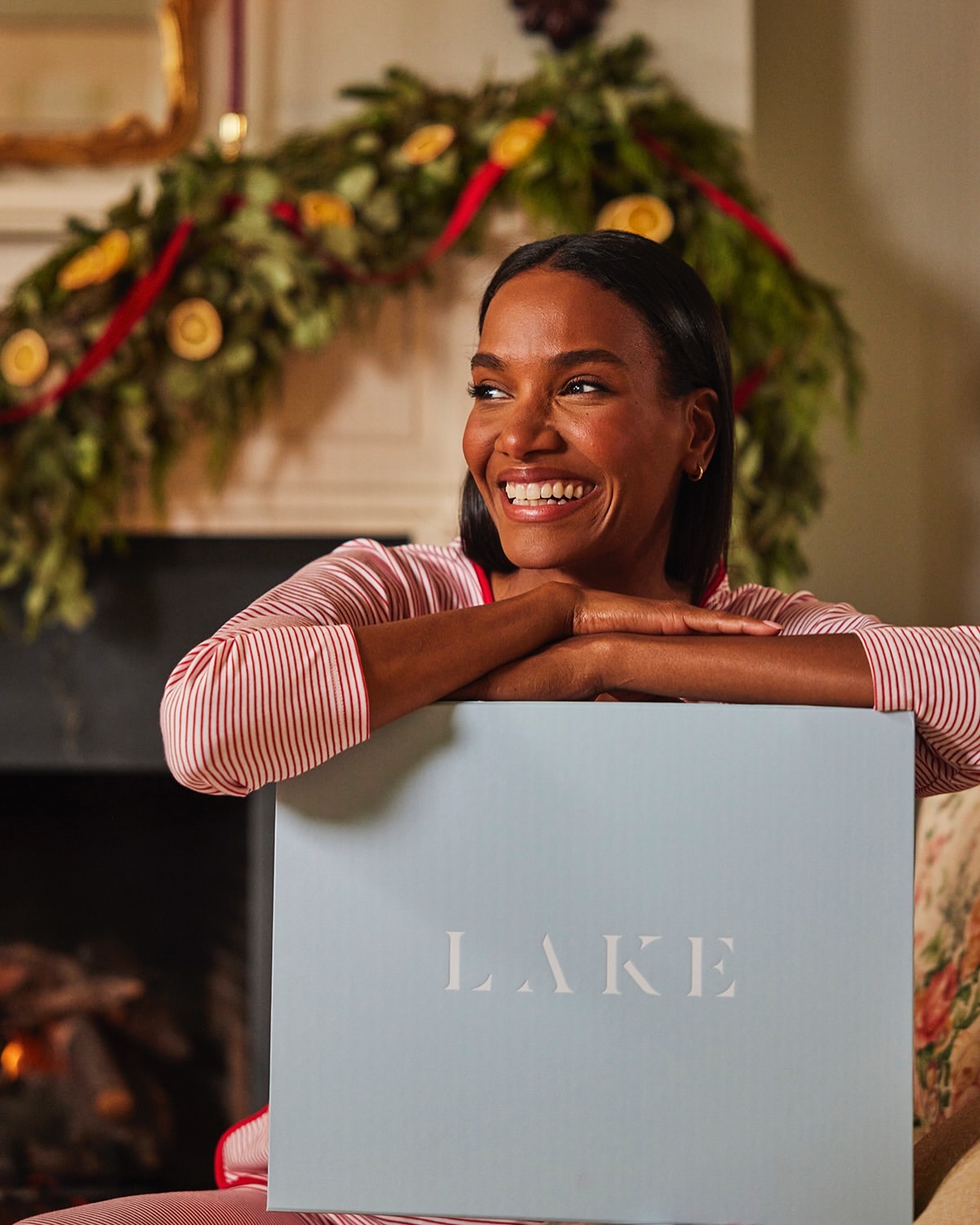 Smiling African-American woman in red and white striped pajamas leaning against a light blue box with the text 'LAKE'. Holiday decor visible.