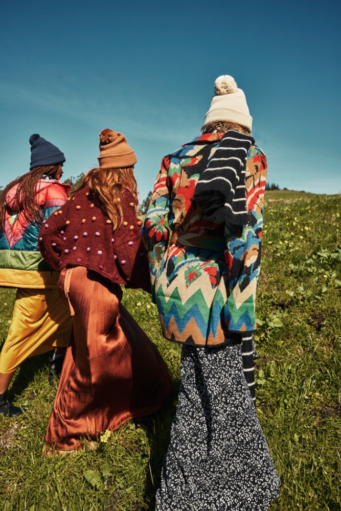 Three models in hats, patterned coats, and skirts standing in a field. The models have their backs turned to the camera on a sunny day.
