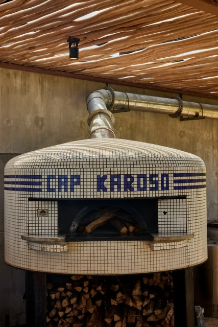 Indoor shot of a tiled pizza oven with wood inside, metal piping above, and a wood slat ceiling in the background.