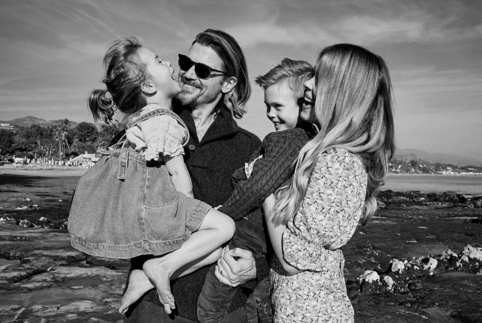 Black and white shot of happy family of four smiling while standing on rocky beach in sunlight, with ocean and mountains visible.