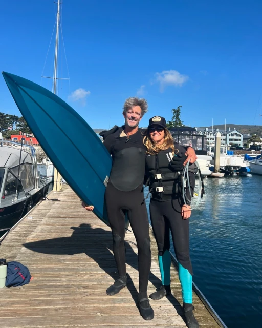 Two people in wetsuits on a dock holding a surfboard, with sailboats and buildings in the background on a bright sunny day.