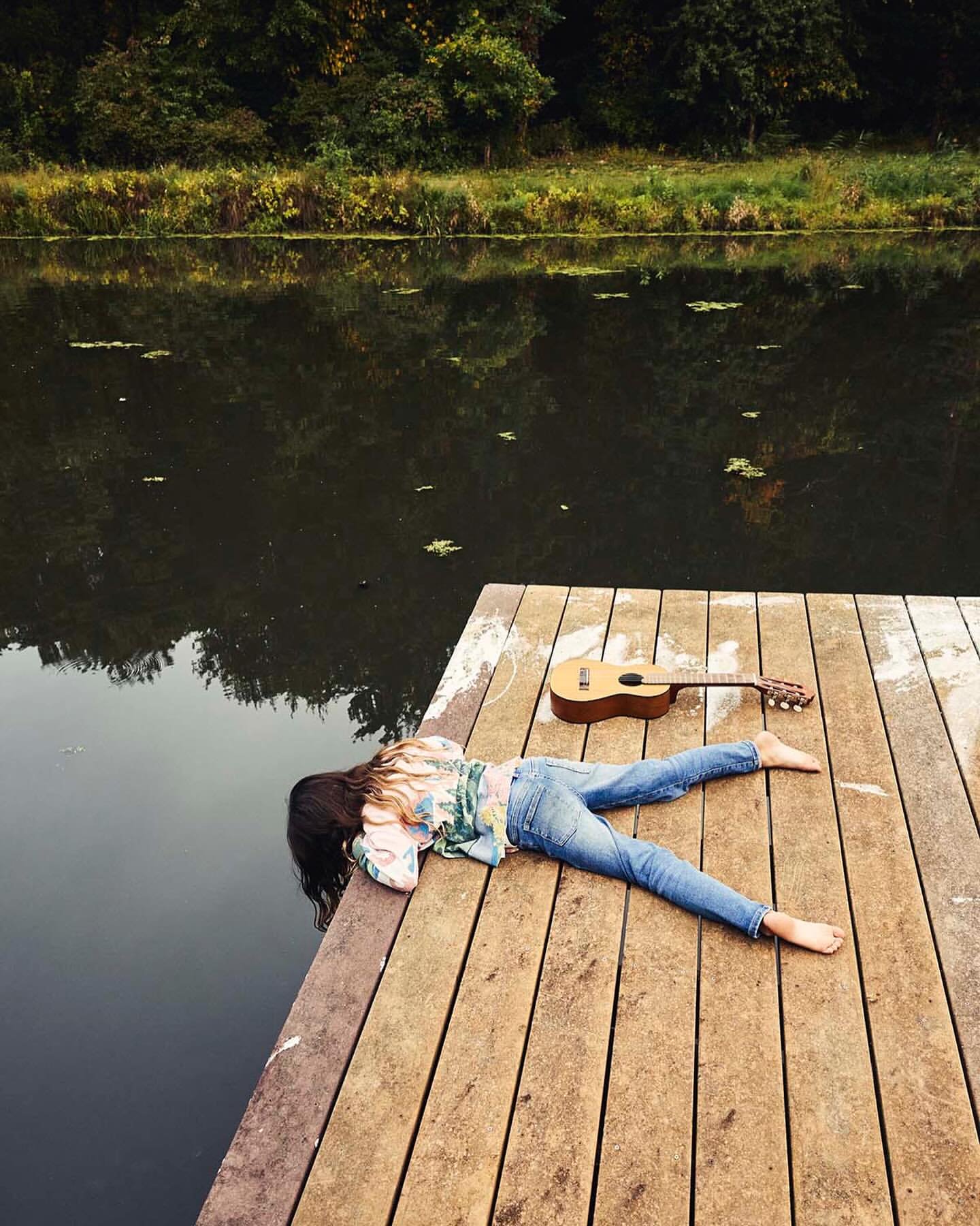Woman laying on a wooden dock next to a calm river with a guitar, surrounded by lush green trees and foliage under an overcast sky.