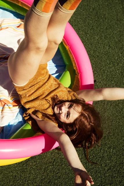 Young red-haired woman in gold top and striped socks lies upside down in colorful inflatable pool on green lawn under natural sunlight.