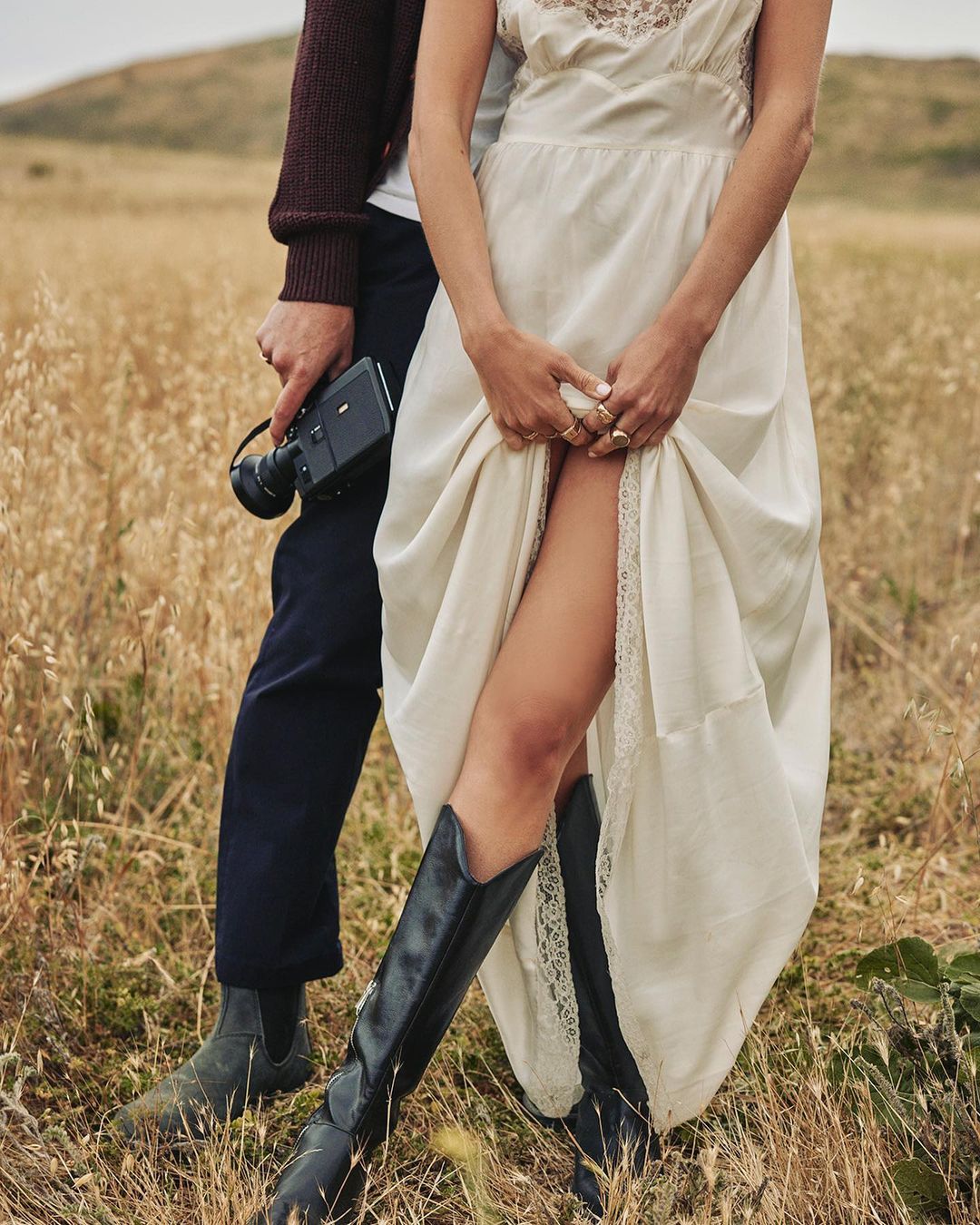 A couple stands in a field of tall grass. The woman wears a white dress and black boots, and the man holds a vintage video camera.