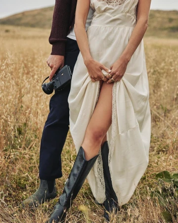 A couple stands in a field of tall grass. The woman wears a white dress and black boots, and the man holds a vintage video camera.