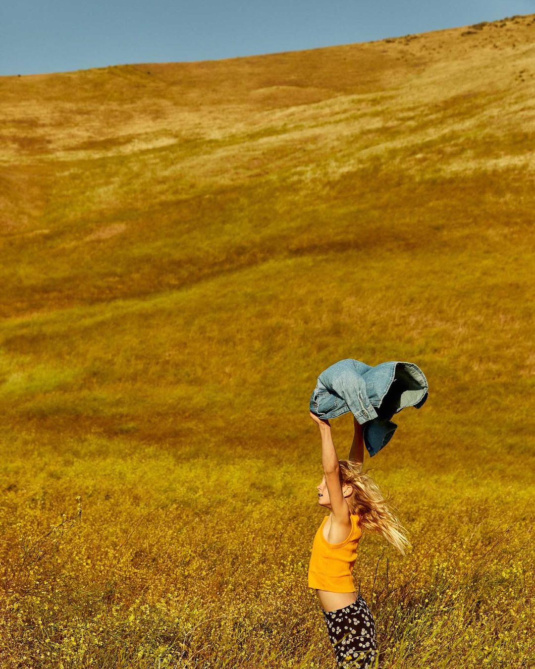 Young girl in yellow top stands in field of wildflowers, holding denim jacket overhead against a background of a golden hillside.