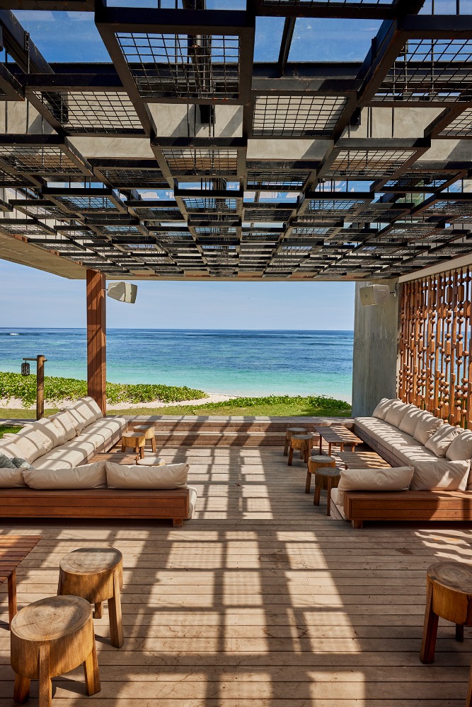 Open-air patio with wooden decking, plush seating, and a patterned overhead trellis casting geometric shadows facing the ocean.