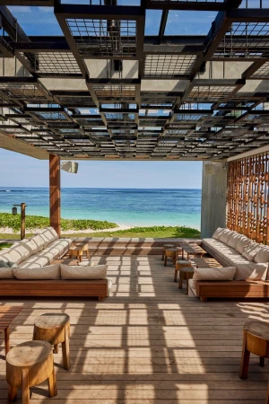Open-air patio with wooden decking, plush seating, and a patterned overhead trellis casting geometric shadows facing the ocean.