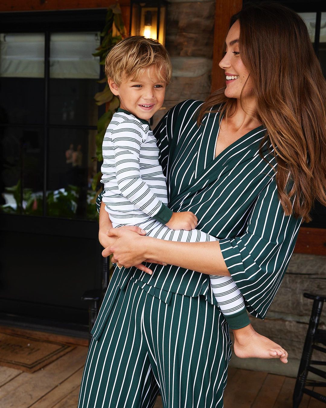 A smiling woman in striped loungewear holds a smiling young boy in matching pajamas near a dark doorway with green foliage.