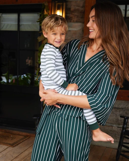 A smiling woman in striped loungewear holds a smiling young boy in matching pajamas near a dark doorway with green foliage.