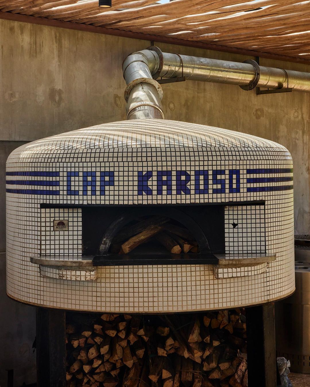 Mosaic tile pizza oven with a metal flue and a stack of firewood in an outdoor setting, under a thatched roof structure.