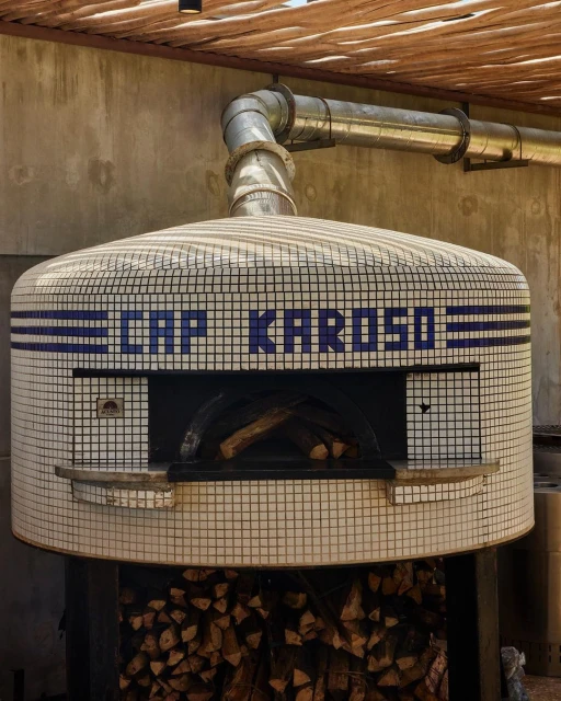 Mosaic tile pizza oven with a metal flue and a stack of firewood in an outdoor setting, under a thatched roof structure.