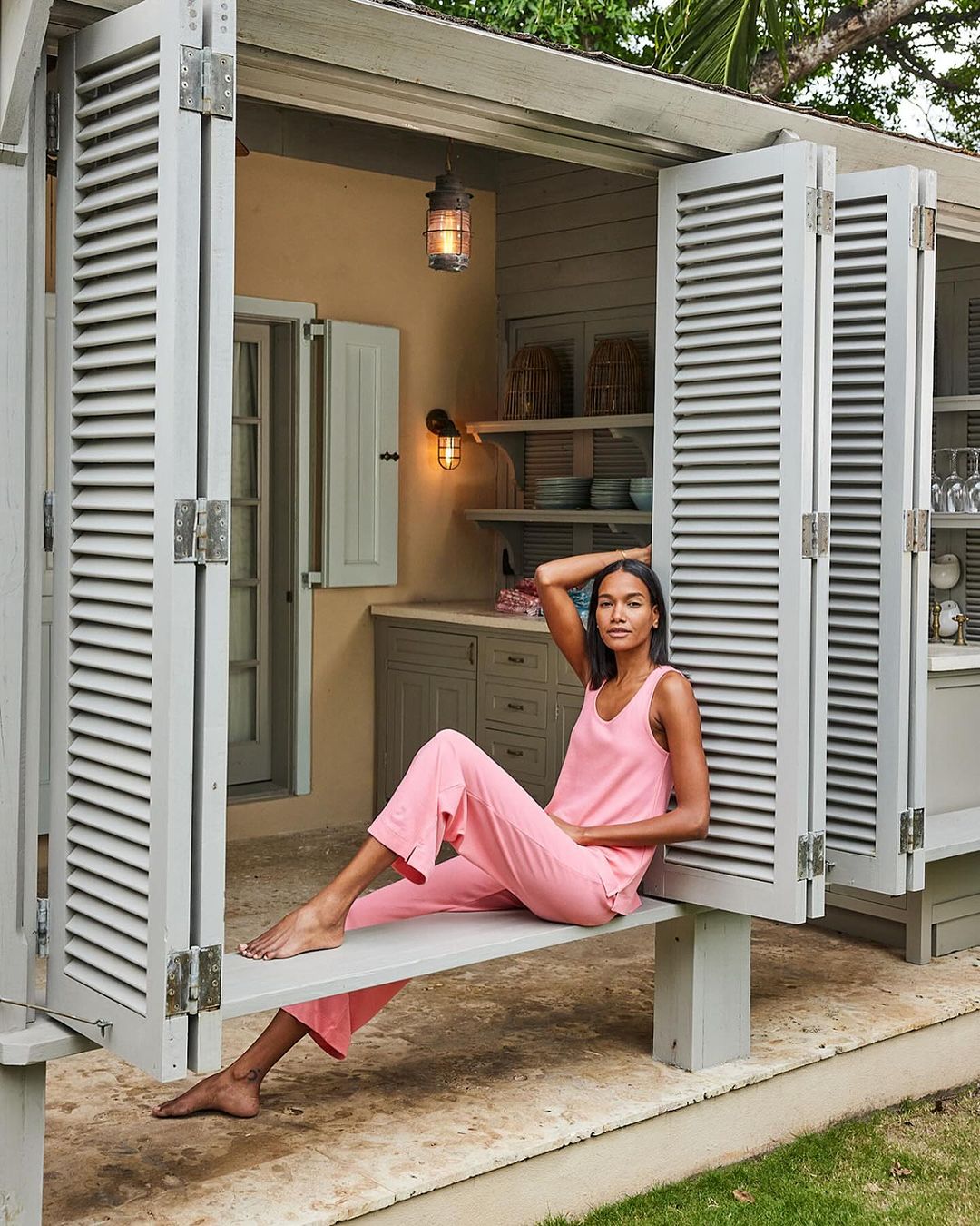 A woman in pink loungewear sits in the open doorway of a neutral-toned building, bathed in natural light. Exterior shutters frame the scene.