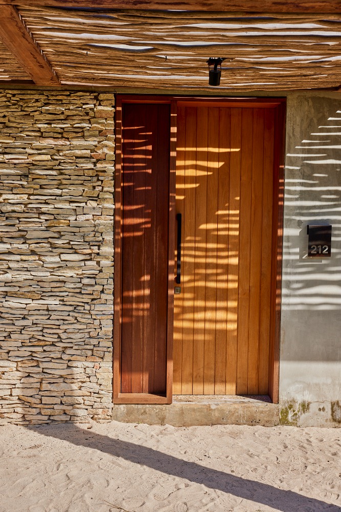 Exterior shot of a modern building with a light wood door, stone wall, and thatched roof casting shadow on the facade during daytime.