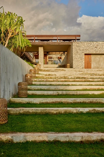 Exterior view of a modern house with concrete stairs and woven light fixtures leading to the entrance, set against a cloudy sky.