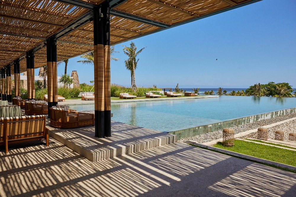 Outdoor lounge area with cushioned seating, a swimming pool, and ocean view under a thatched roof pergola on a sunny day.