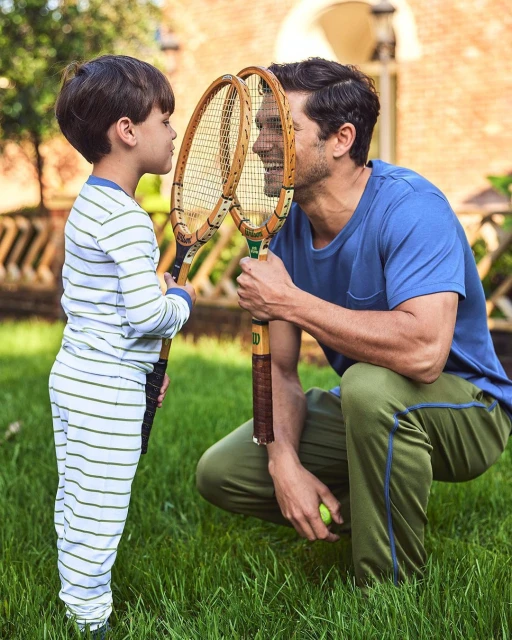 A dad and his son playing outside with tennis rackets, both are wearing matching pajamas. The father is crouching and smiling at his son.