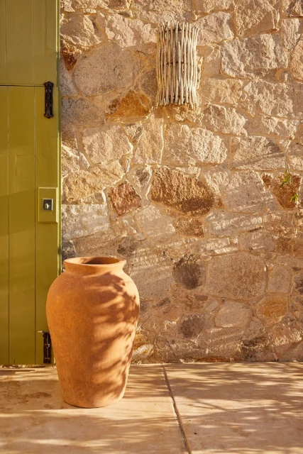 Earthenware pot stands on a stone surface beside a chartreuse door set against a sunlit stone wall in an outdoor setting.