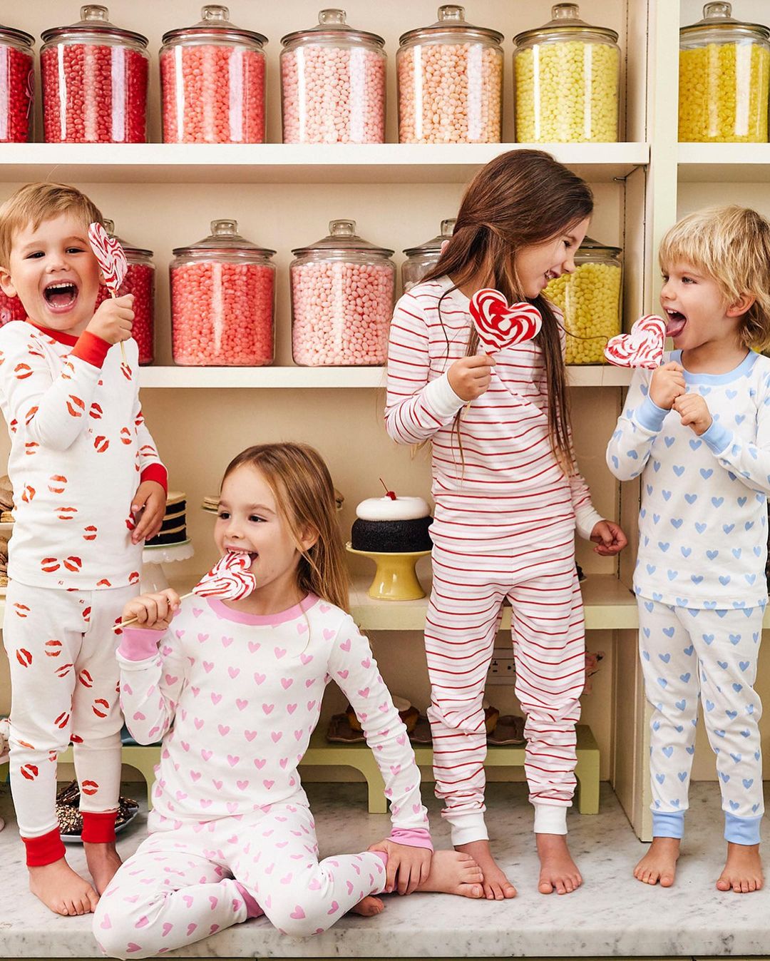 Four smiling children in patterned pajamas are indoors with lollipops. They are in front of shelves filled with jars of candy.