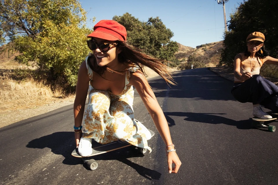 Two women skateboard on asphalt road surrounded by trees. One wears a red hat and floral jumpsuit, smiling in sunny weather.