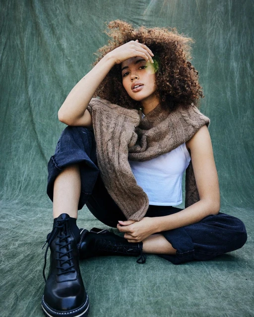 Woman with afro poses in jeans, black boots, and brown shawl against a mottled green backdrop, creating a stylish, casual fashion portrait.