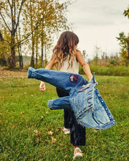 A young girl with long brown hair walks through a grassy field, carrying a denim jacket with decorative patches on her back.