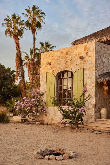 Exterior of a stone building with arched window, green shutters, and palm trees set in a sandy landscape under a blue sky.