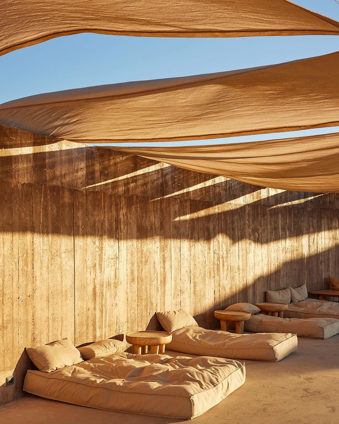 Outdoor lounge area with fabric shade structure, featuring cushioned lounge platforms and small wooden tables against a textured wall.