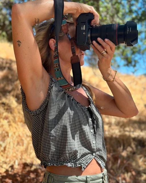 Woman in gingham top takes photo in outdoor setting with professional DSLR camera. Shoulder-length blonde hair. Turquoise and brown strap.