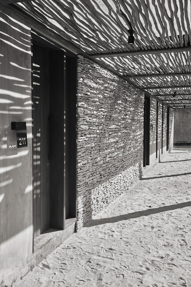 Exterior of a modern building with stone walls, a shaded walkway, and sunlight creating dappled shadows on the sandy ground.