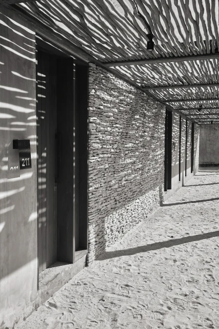 Exterior of a modern building with stone walls, a shaded walkway, and sunlight creating dappled shadows on the sandy ground.
