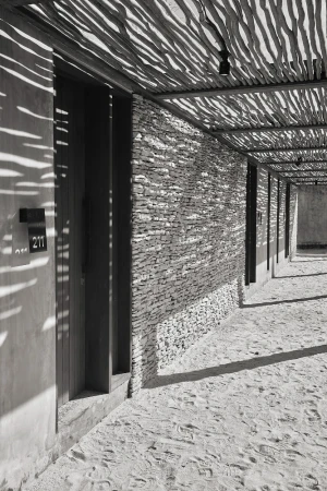 Exterior of a modern building with stone walls, a shaded walkway, and sunlight creating dappled shadows on the sandy ground.