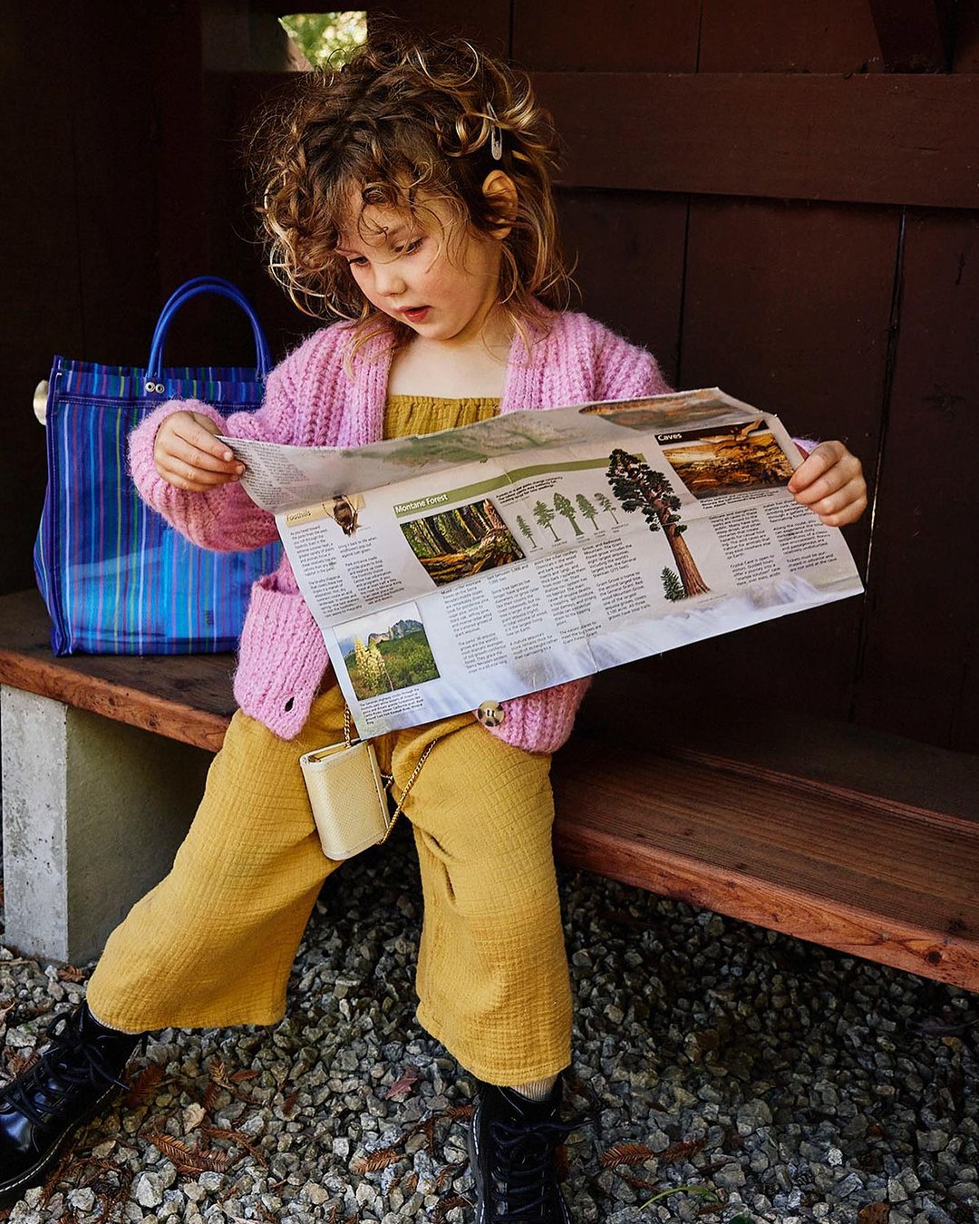 Young girl in pink cardigan and yellow pants sits on a bench outdoors, studying a nature map with a blue bag beside her.