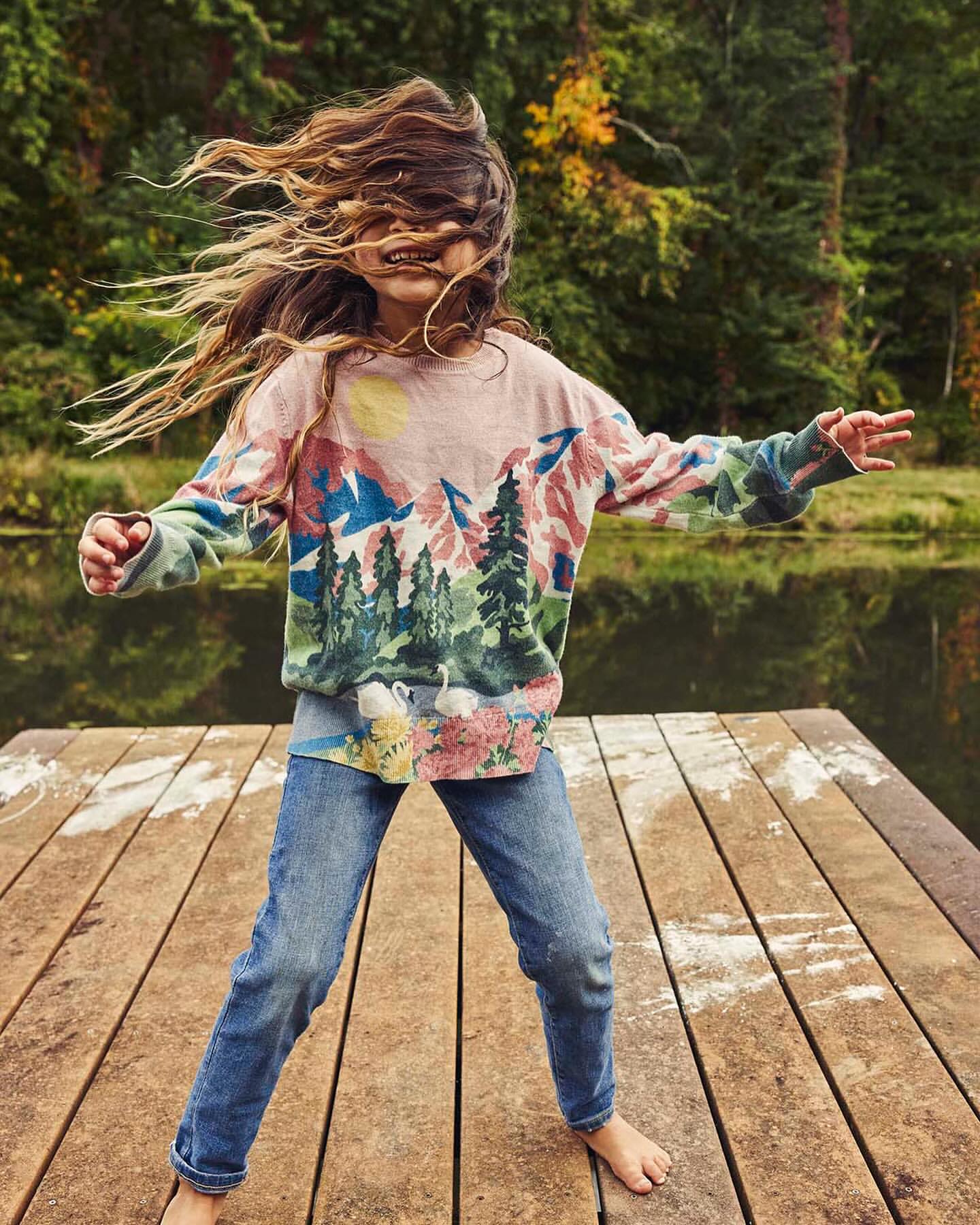 A young girl with long brown hair stands on a wooden dock next to a lake, wearing a mountain-themed shirt and jeans, smiling.